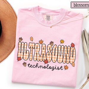 Puede incluir: Camiseta rosa claro con la frase "ULTRASOUND technologist" en una fuente retro. La palabra "ULTRASOUND" tiene un patr&oacute;n de cuadros con hojas de oto&ntilde;o y bellotas.