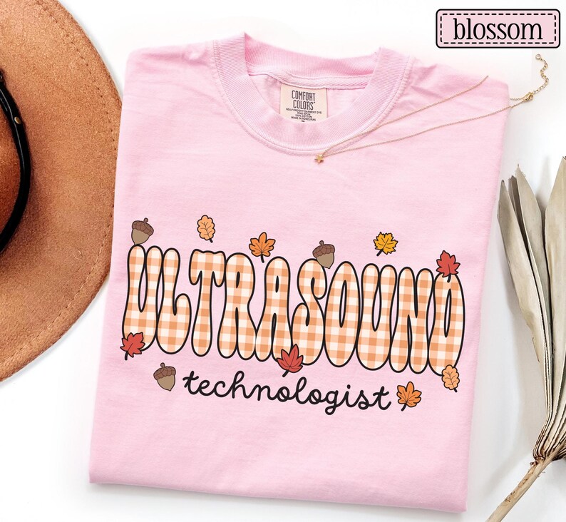 Puede incluir: Camiseta rosa claro con la frase "ULTRASOUND technologist" en una fuente retro. La palabra "ULTRASOUND" tiene un patr&oacute;n de cuadros con hojas de oto&ntilde;o y bellotas.