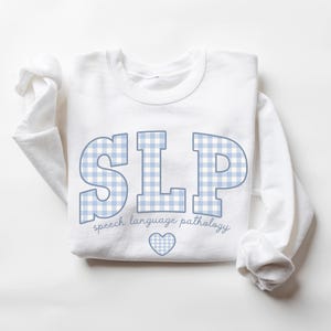 May include: White sweatshirt with a blue and white checkered design that spells out "SLP" and the text "speech language pathology" below. A small blue heart is centered below the text.
