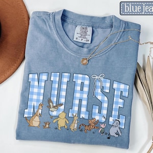 K&ouml;nnte beinhalten: Ein blaues Comfort Colors T-Shirt mit dem Wort "NURSE" in gro&szlig;en, hellblauen Vichy-Buchstaben. Unter dem Wort befinden sich Zeichentrickfiguren. Eine goldene Halskette mit Anh&auml;nger ist &uuml;ber dem Shirt drapiert.