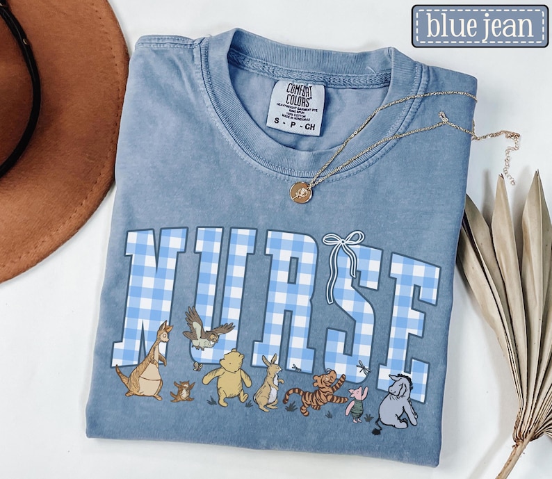 K&ouml;nnte beinhalten: Ein blaues Comfort Colors T-Shirt mit dem Wort "NURSE" in gro&szlig;en, hellblauen Vichy-Buchstaben. Unter dem Wort befinden sich Zeichentrickfiguren. Eine goldene Halskette mit Anh&auml;nger ist &uuml;ber dem Shirt drapiert.