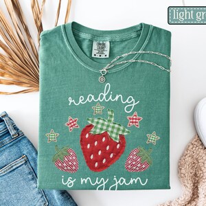May include: A light green t-shirt featuring a graphic of three strawberries and stars with the text reading is my jam. The shirt has a crew neckline and a Comfort Colors brand label on the inside collar. A silver chain necklace rests on the shirt.