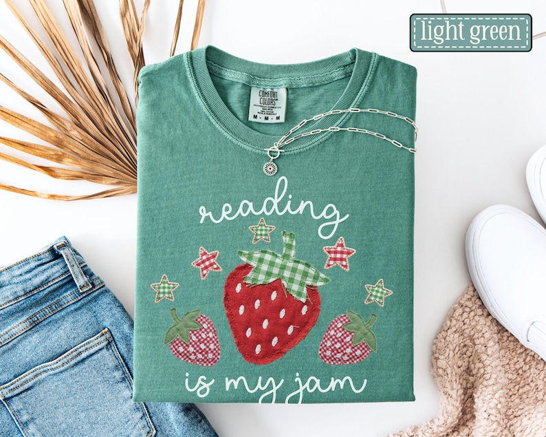 May include: A light green t-shirt featuring a graphic of three strawberries and stars with the text reading is my jam. The shirt has a crew neckline and a Comfort Colors brand label on the inside collar. A silver chain necklace rests on the shirt.