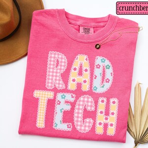 May include: A bright pink t-shirt with the words "RAD TECH" in a patchwork design. The letters are decorated with gingham, floral, and scalloped patterns. A gold necklace and a brown hat are also visible.