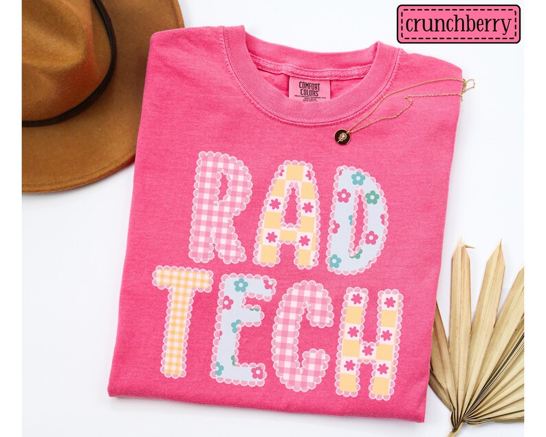 May include: A bright pink t-shirt with the words "RAD TECH" in a patchwork design. The letters are decorated with gingham, floral, and scalloped patterns. A gold necklace and a brown hat are also visible.