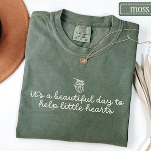 May include: A moss green t-shirt with the text "it's a beautiful day to help little hearts" in white script. The shirt has a crew neck and short sleeves. A gold necklace with a small pendant is draped on the shirt.