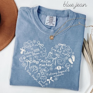 May include: Blue jean Comfort Colors t-shirt with a white heart-shaped graphic. The graphic includes baby-related illustrations and the words "NICU nurse." A gold necklace with a pendant is draped across the shirt. The shirt is folded on a white surface.