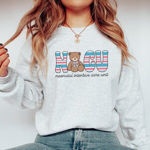 Puede incluir: Una sudadera gris con un gr&aacute;fico colorido de un osito de peluche y el texto "NICU unidad de cuidados intensivos neonatales".