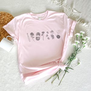May include: A pink t-shirt with a black graphic of a crochet hook, yarn, and flowers. The graphic says "Crafty AF".