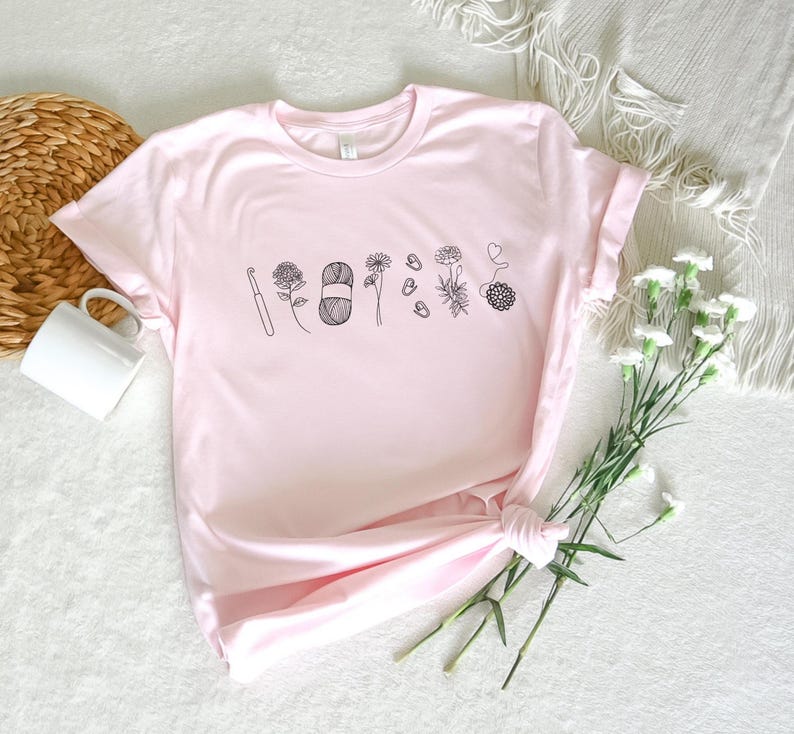 May include: A pink t-shirt with a black graphic of a crochet hook, yarn, and flowers. The graphic says "Crafty AF".