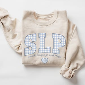 May include: A beige sweatshirt with the text "SLP" in blue and white gingham print. The text "speech language pathology" is written below the letters. A small heart is below the text.