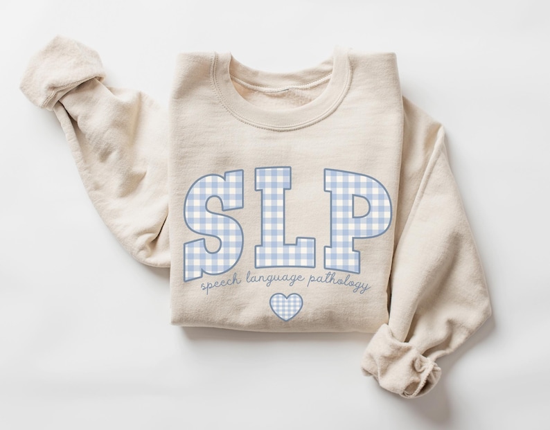 May include: A beige sweatshirt with the text "SLP" in blue and white gingham print. The text "speech language pathology" is written below the letters. A small heart is below the text.