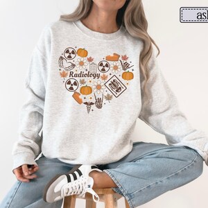 May include: An ash gray crewneck sweatshirt featuring a heart-shaped graphic on the chest. The design includes the word Radiology, pumpkins, flowers, autumn leaves, radiation symbols, a skeleton hand, and a medical caduceus in orange and brown.