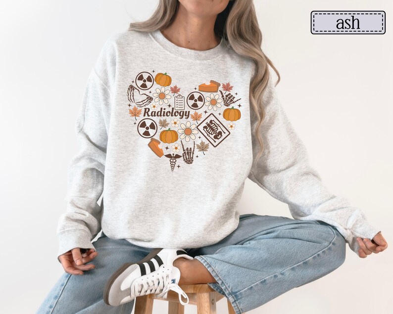 May include: An ash gray crewneck sweatshirt featuring a heart-shaped graphic on the chest. The design includes the word Radiology, pumpkins, flowers, autumn leaves, radiation symbols, a skeleton hand, and a medical caduceus in orange and brown.