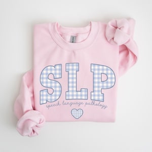 May include: Pink sweatshirt with a blue and white gingham pattern spelling "SLP" and the text "speech language pathology" below. A small heart is centered below the text.