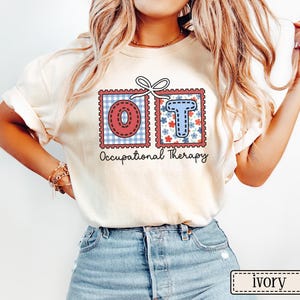 May include: Ivory-colored t-shirt featuring the letters "OT" in decorative postage stamp designs. The "O" is red with a blue gingham background, and the "T" is blue with a floral background. The words "Occupational Therapy" are printed below.