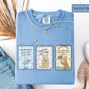 May include: Light blue "washed denim" Comfort Colors t-shirt with Winnie the Pooh characters and the phrases "helping you grow your own way", "Speech Therapy", and "Communication looks different for everyone". A silver necklace is draped on the shirt.