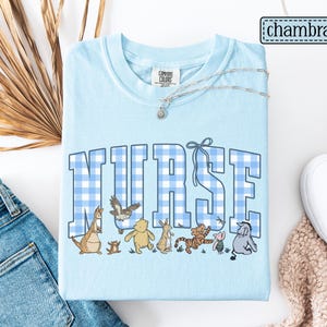 K&ouml;nnte beinhalten: Hellblaues T-Shirt mit dem Wort "NURSE" in gro&szlig;en, blau-wei&szlig; karierten Buchstaben. Darunter befinden sich Zeichentrickfiguren. Das Shirt hat ein Comfort Colors-Etikett und eine silberne Halskette. Das Wort "chambray" befindet sich oben rechts.