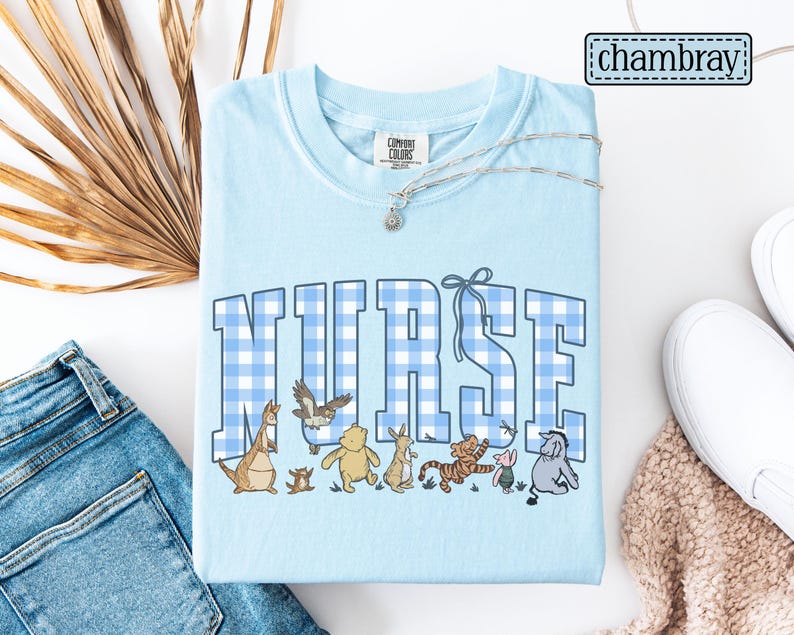K&ouml;nnte beinhalten: Hellblaues T-Shirt mit dem Wort "NURSE" in gro&szlig;en, blau-wei&szlig; karierten Buchstaben. Darunter befinden sich Zeichentrickfiguren. Das Shirt hat ein Comfort Colors-Etikett und eine silberne Halskette. Das Wort "chambray" befindet sich oben rechts.