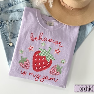 K&ouml;nnte beinhalten: Ein hellviolettes T-Shirt mit einer Grafik aus drei Erdbeeren mit rot-gr&uuml;nem Vichy-Muster. Der Text "behavior is my jam" ist in roter Schreibschrift gedruckt, begleitet von kleinen rot-gr&uuml;nen Vichy-Sternen, die die Fr&uuml;chte umgeben.