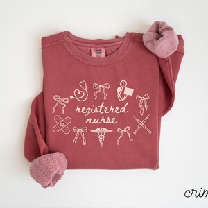 May include: Crimson-colored sweatshirt with the words "registered nurse" and medical-related graphics in white. The design includes a stethoscope, band-aid, and syringes. The sleeves have a soft, pinkish-red lining.