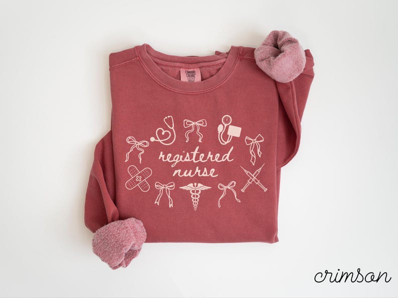 May include: Crimson-colored sweatshirt with the words "registered nurse" and medical-related graphics in white. The design includes a stethoscope, band-aid, and syringes. The sleeves have a soft, pinkish-red lining.