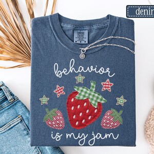 K&ouml;nnte beinhalten: Ein kurz&auml;rmeliges T-Shirt in Denim-Blau mit einer Grafik aus drei Erdbeeren mit rot-gr&uuml;nem Karomuster. Der Text "behavior is my jam" ist in wei&szlig;er Schreibschrift gedruckt. Kleine rot-gr&uuml;ne Karosterne umgeben die Erdbeeren.