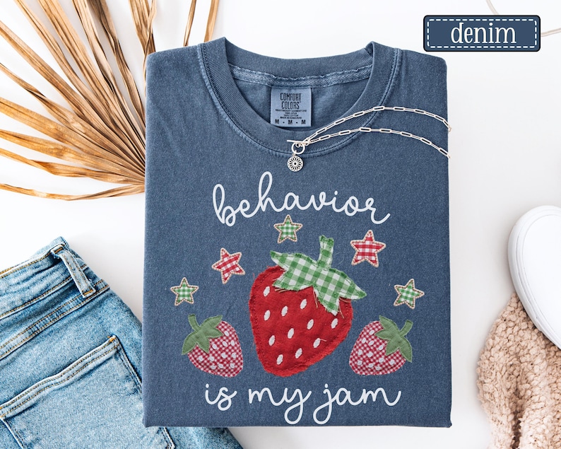 K&ouml;nnte beinhalten: Ein kurz&auml;rmeliges T-Shirt in Denim-Blau mit einer Grafik aus drei Erdbeeren mit rot-gr&uuml;nem Karomuster. Der Text "behavior is my jam" ist in wei&szlig;er Schreibschrift gedruckt. Kleine rot-gr&uuml;ne Karosterne umgeben die Erdbeeren.