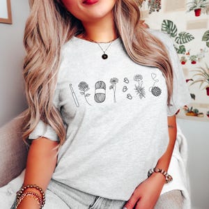 May include: A gray t-shirt with a black line drawing of a crochet hook, yarn, flowers, and other crochet supplies. The shirt has a crew neck and short sleeves.