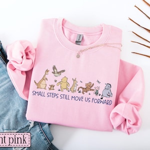 May include: Pale pink sweatshirt with a crew neck and long sleeves. The front displays a cartoon graphic and the text "SMALL STEPS STILL MOVE US FORWARD". A necklace and blue jeans are also visible.