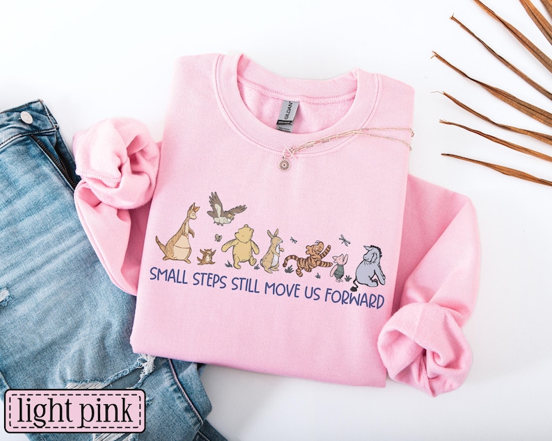 May include: Pale pink sweatshirt with a crew neck and long sleeves. The front displays a cartoon graphic and the text "SMALL STEPS STILL MOVE US FORWARD". A necklace and blue jeans are also visible.