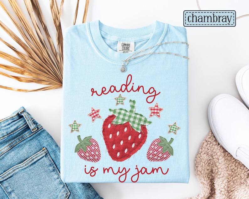 May include: A light blue t-shirt featuring a graphic of three strawberries with red and white chequered patterns and green chequered leaves. The text "reading is my jam" is printed in red script, accompanied by small red and green chequered stars.