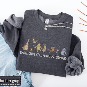 May include: Dark grey sweatshirt featuring a cartoon character graphic and the text "SMALL STEPS STILL MOVE US FORWARD." A silver necklace with a pendant is draped across the sweatshirt. The sweatshirt is folded next to blue denim jeans.