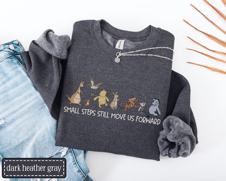 May include: Dark grey sweatshirt featuring a cartoon character graphic and the text "SMALL STEPS STILL MOVE US FORWARD." A silver necklace with a pendant is draped across the sweatshirt. The sweatshirt is folded next to blue denim jeans.