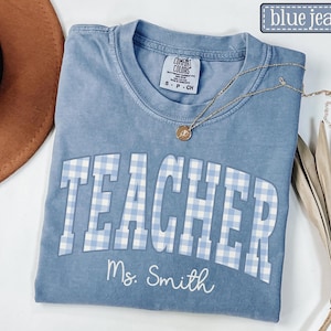 Puede incluir: Una camiseta azul vaquera Comfort Colors con la palabra "TEACHER" en estampado de cuadros blancos y celeste. El nombre "Ms. Smith" está impreso debajo de la palabra "TEACHER". Un collar dorado con un pequeño colgante está sobre la camisa.