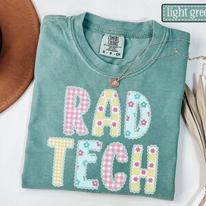 May include: Light green t-shirt with the words "RAD TECH" in a colorful, patterned design. The letters are outlined with a scalloped edge and filled with gingham and floral patterns. A gold necklace with a pendant is draped across the shirt.