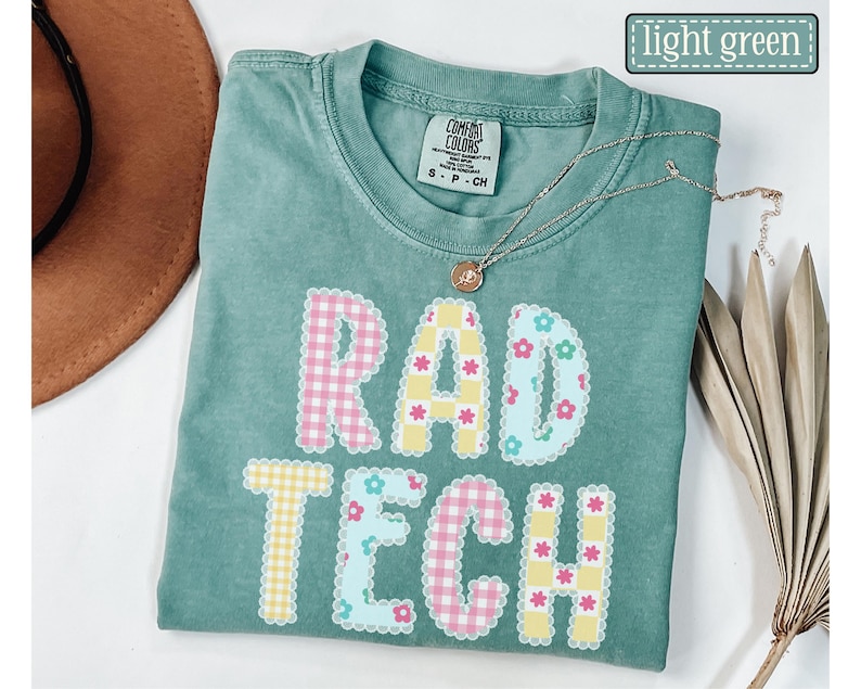 May include: Light green t-shirt with the words "RAD TECH" in a colorful, patterned design. The letters are outlined with a scalloped edge and filled with gingham and floral patterns. A gold necklace with a pendant is draped across the shirt.