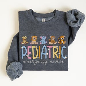 May include: A dark gray sweatshirt with the text "PEDIATRIC emergency nurse" in colorful letters with flowers. The text is surrounded by five cartoon animals: a giraffe, a lion, an elephant, a monkey, and a tiger.