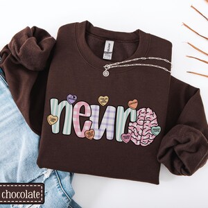 May include: A dark chocolate brown sweatshirt with the word "neuro" in a colorful design, featuring a brain graphic and candy hearts. The sweatshirt is paired with blue jeans and a silver necklace.