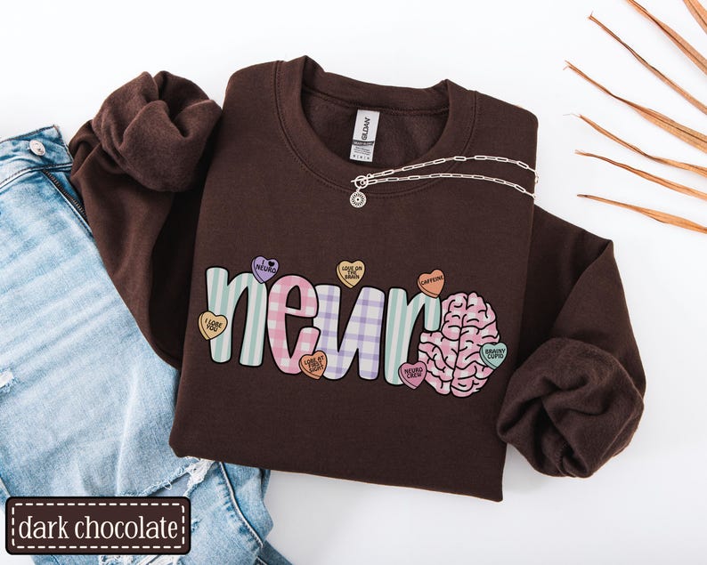 May include: A dark chocolate brown sweatshirt with the word "neuro" in a colorful design, featuring a brain graphic and candy hearts. The sweatshirt is paired with blue jeans and a silver necklace.
