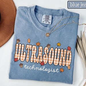 Puede incluir: Una camiseta azul vaquera Comfort Colors con las palabras "ULTRASOUND technologist" en una fuente retro con un patr&oacute;n de cuadros. La camiseta est&aacute; decorada con hojas de oto&ntilde;o y bellotas, y un collar dorado.