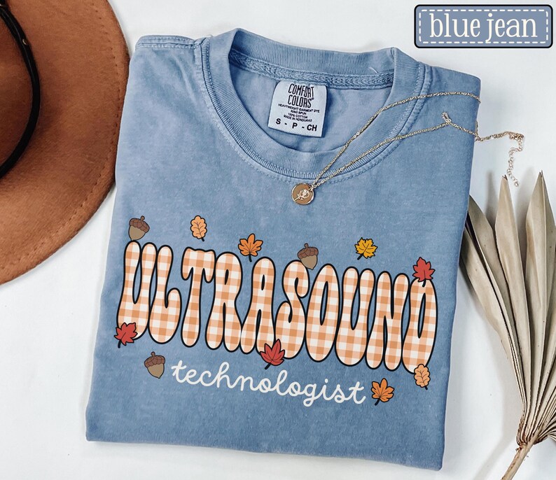 Puede incluir: Una camiseta azul vaquera Comfort Colors con las palabras "ULTRASOUND technologist" en una fuente retro con un patr&oacute;n de cuadros. La camiseta est&aacute; decorada con hojas de oto&ntilde;o y bellotas, y un collar dorado.