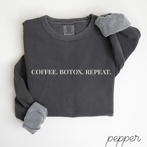May include: Dark gray sweatshirt with the text "COFFEE. BOTOX. REPEAT." in white. The sweatshirt has a crew neck and long sleeves with gray fleece cuffs. The word "pepper" is written in cursive in the bottom right corner.