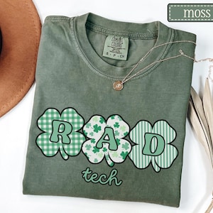May include: Moss green t-shirt with a clover design spelling out "RAD" and the word "tech" below. The clover designs have different patterns, including gingham, shamrocks, and stripes. A gold necklace rests on the shirt.