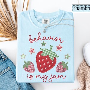 K&ouml;nnte beinhalten: Ein hellblaues T-Shirt mit einer Grafik aus drei roten Erdbeeren mit gr&uuml;n karierten Bl&auml;ttern und kleinen karierten Sternen. Der Text "behavior is my jam" ist in roter Schreibschrift aufgedruckt. Das T-Shirt hat einen Rundhalsausschnitt.