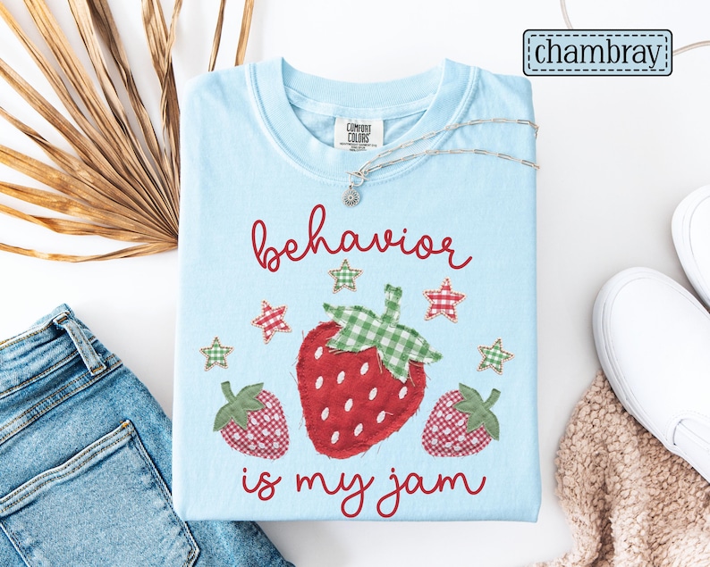 K&ouml;nnte beinhalten: Ein hellblaues T-Shirt mit einer Grafik aus drei roten Erdbeeren mit gr&uuml;n karierten Bl&auml;ttern und kleinen karierten Sternen. Der Text "behavior is my jam" ist in roter Schreibschrift aufgedruckt. Das T-Shirt hat einen Rundhalsausschnitt.