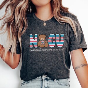Puede incluir: Una camiseta gris oscuro con un gr&aacute;fico de las letras "NCU" en rayas rosas, azules y blancas. Un oso de peluche marr&oacute;n con un lazo azul est&aacute; en el centro de las letras. El texto "neonatal intensive care unit" est&aacute; debajo del gr&aacute;fico.