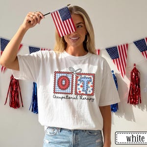 May include: White t-shirt with a graphic design featuring the letters "OT" in red and blue postage stamp style boxes. The shirt also has the words "Occupational Therapy" printed below the letters. The person is holding a small American flag.