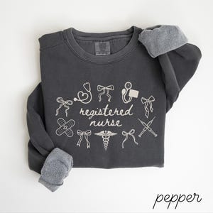 May include: A dark gray sweatshirt with the words "registered nurse" and medical-themed graphics in white. The design includes a stethoscope, band-aid, and syringe. The sleeves have gray, fuzzy cuffs. The word "pepper" is written in cursive at the bottom.