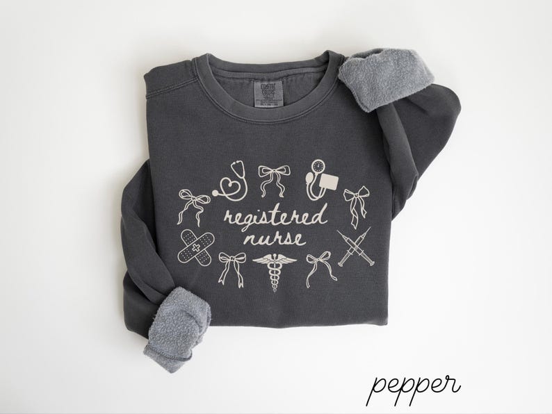 May include: A dark gray sweatshirt with the words "registered nurse" and medical-themed graphics in white. The design includes a stethoscope, band-aid, and syringe. The sleeves have gray, fuzzy cuffs. The word "pepper" is written in cursive at the bottom.